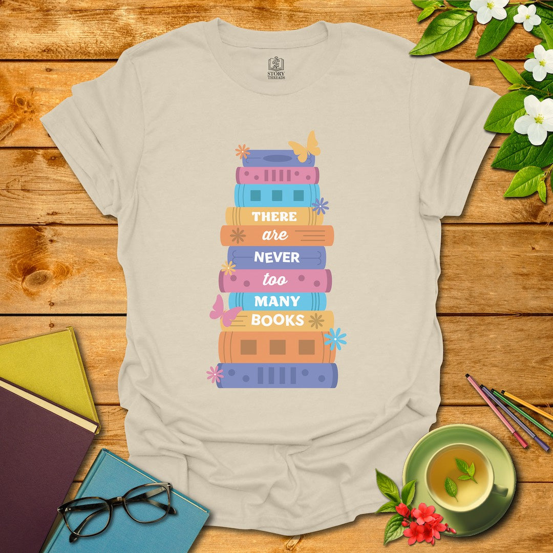 Natural unisex T-shirt with the saying 'There are never too many books' on a stack of colorful books, surrounded by butterflies and flowers, against a backdrop of wooden planks, books, and a cup of tea.
