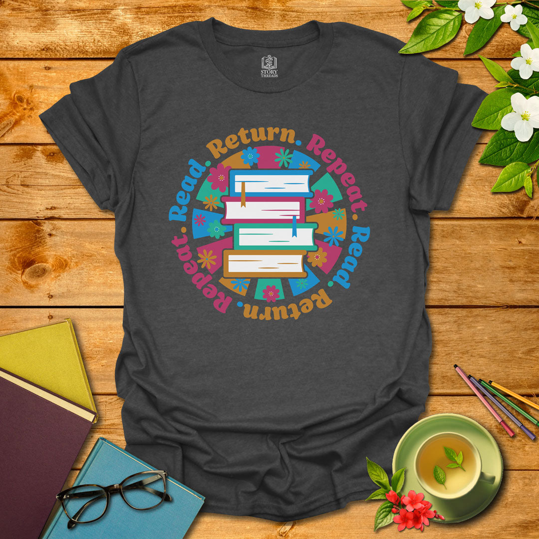 Dark heather unisex T-shirt with a colorful book stack and floral design, encircled by the phrase 'Read. Return. Repeat.' Displayed on a styled background with books and a cup of tea.