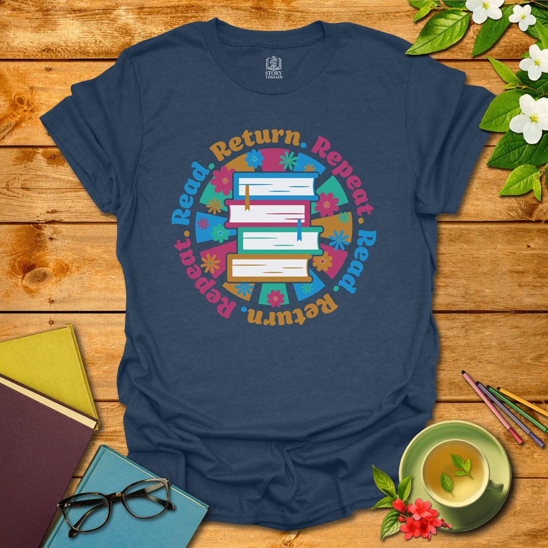 Heather navy unisex T-shirt with a colorful book stack and floral design, encircled by the phrase 'Read. Return. Repeat.' Displayed on a styled background with books and a cup of tea.