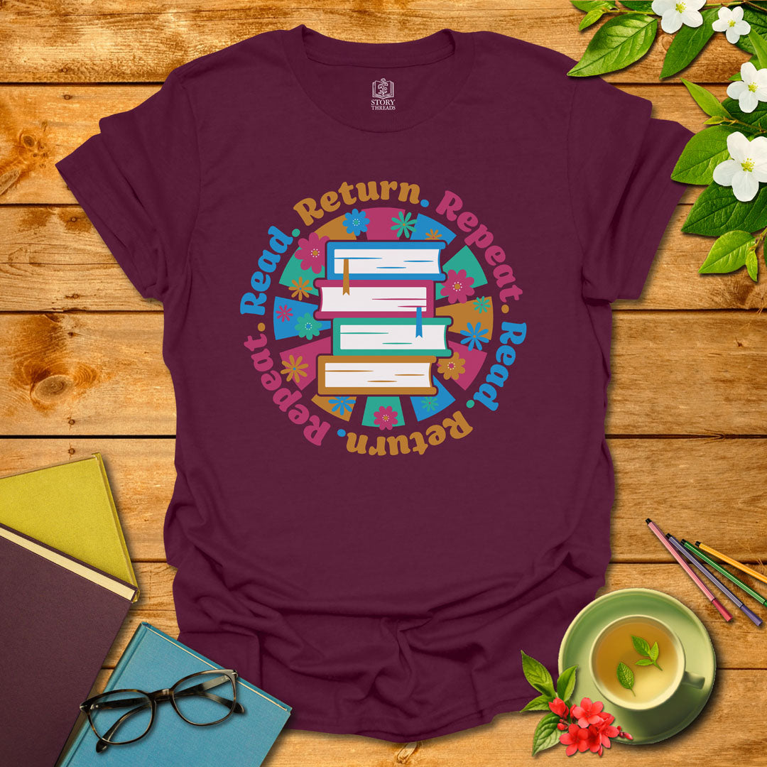 Maroon unisex T-shirt with a colorful book stack and floral design, encircled by the phrase 'Read. Return. Repeat.' Displayed on a styled background with books and a cup of tea.