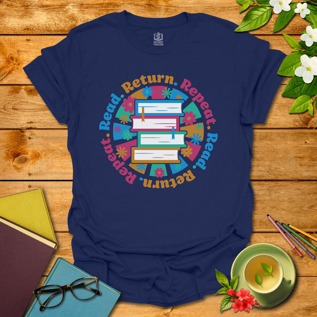 Navy unisex T-shirt with a colorful book stack and floral design, encircled by the phrase 'Read. Return. Repeat.' Displayed on a styled background with books and a cup of tea.