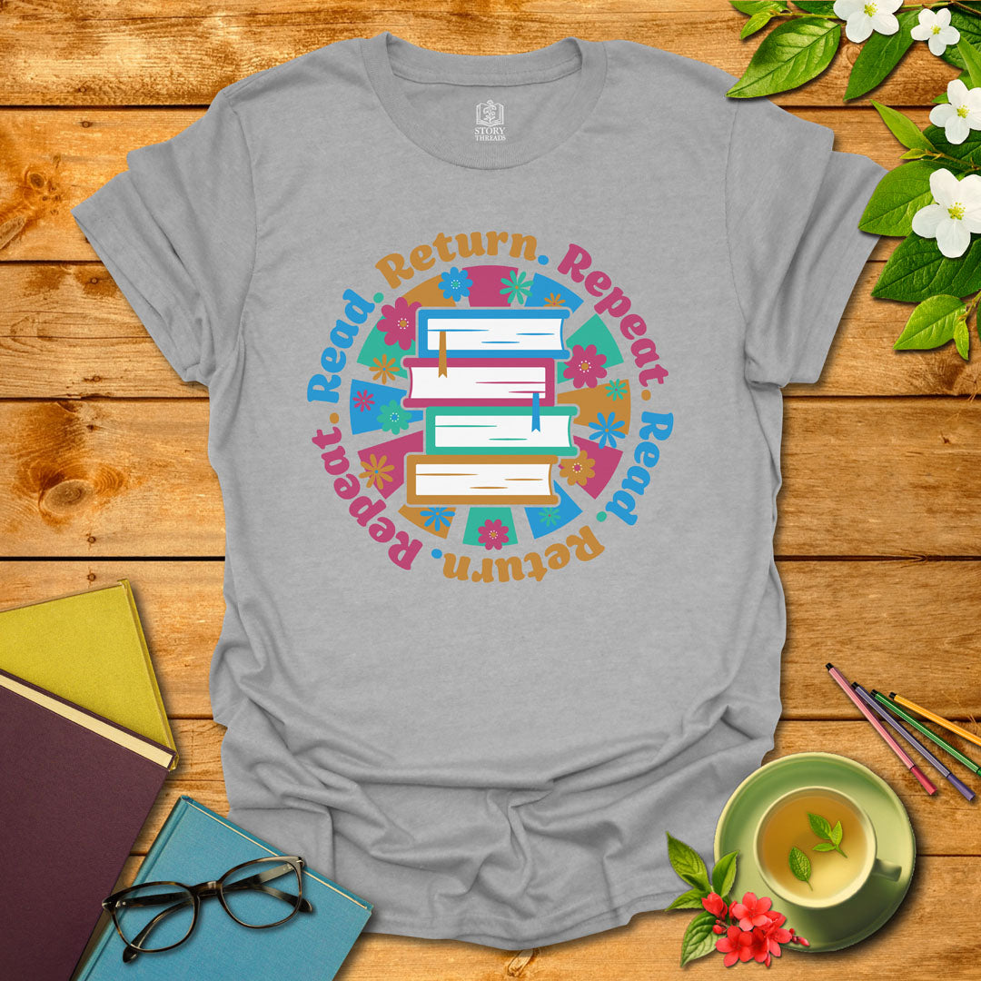 Sport grey unisex T-shirt with a colorful book stack and floral design, encircled by the phrase 'Read. Return. Repeat.' Displayed on a styled background with books and a cup of tea.