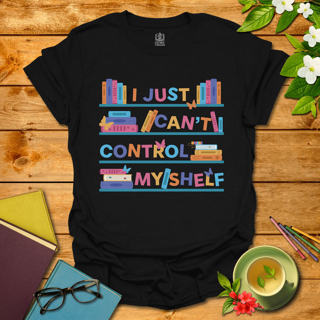 Black unisex T-shirt featuring a colorful bookshelf design with butterflies and the integrated pun 'I Just Can't Control Myshelf', displayed on wooden planks with books and a cup of tea.