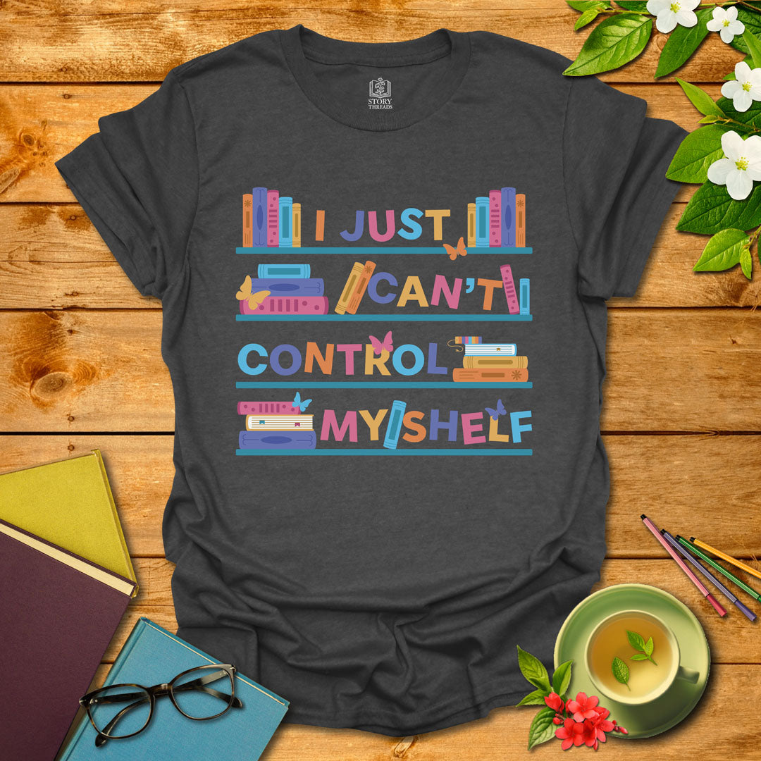 Dark heather unisex T-shirt featuring a colorful bookshelf design with butterflies and the integrated pun 'I Just Can't Control Myshelf', displayed on wooden planks with books and a cup of tea.