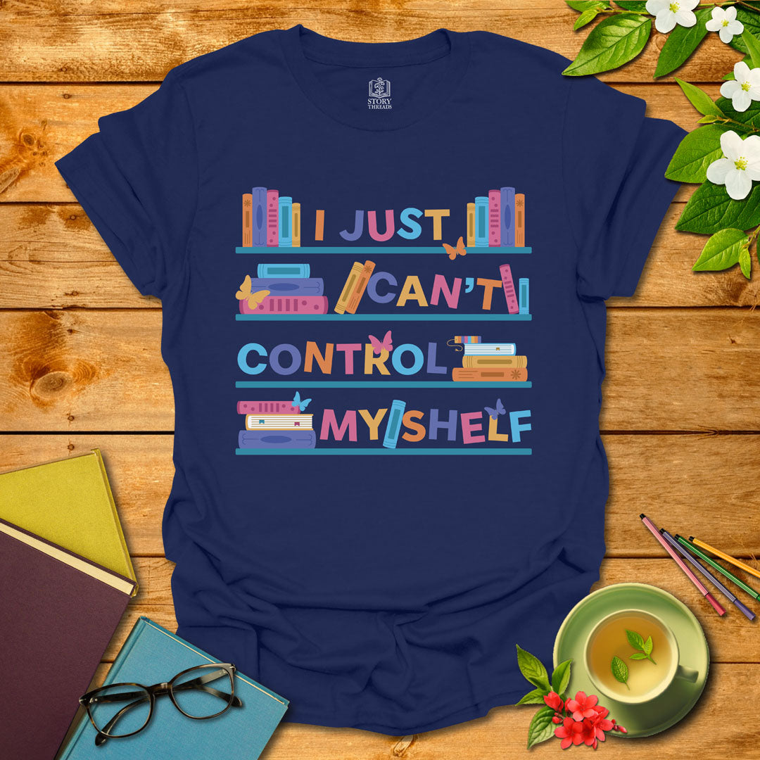 Navy unisex T-shirt featuring a colorful bookshelf design with butterflies and the integrated pun 'I Just Can't Control Myshelf', displayed on wooden planks with books and a cup of tea.