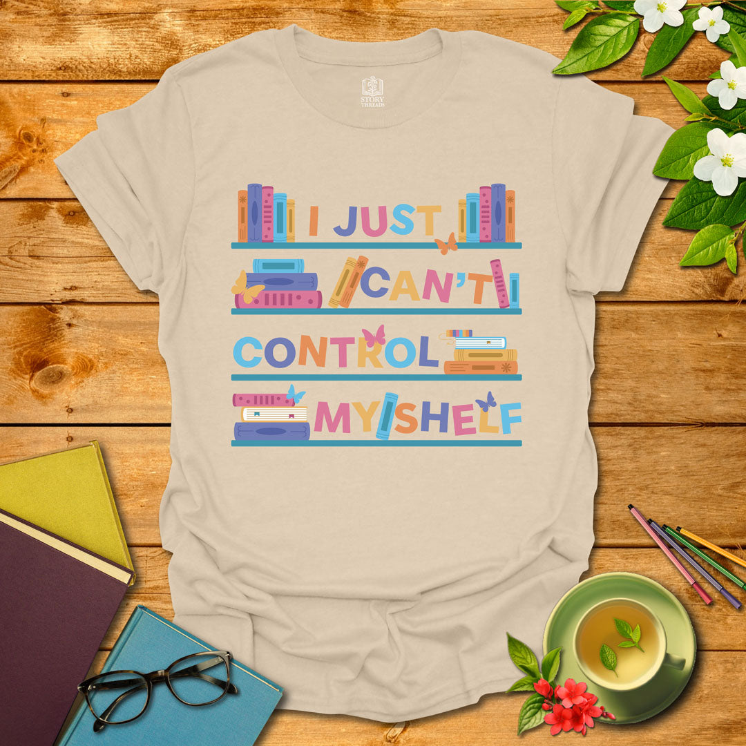 Sand unisex T-shirt featuring a colorful bookshelf design with butterflies and the integrated pun 'I Just Can't Control Myshelf', displayed on wooden planks with books and a cup of tea.