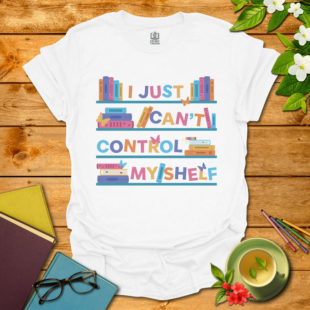 White unisex T-shirt featuring a colorful bookshelf design with butterflies and the integrated pun 'I Just Can't Control Myshelf', displayed on wooden planks with books and a cup of tea.