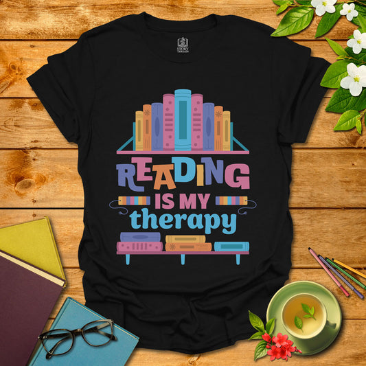 Black unisex T-shirt with 'Reading Is My Therapy' in colorful letters among books on a floating shelf, presented on a styled wooden background with a cup of tea.